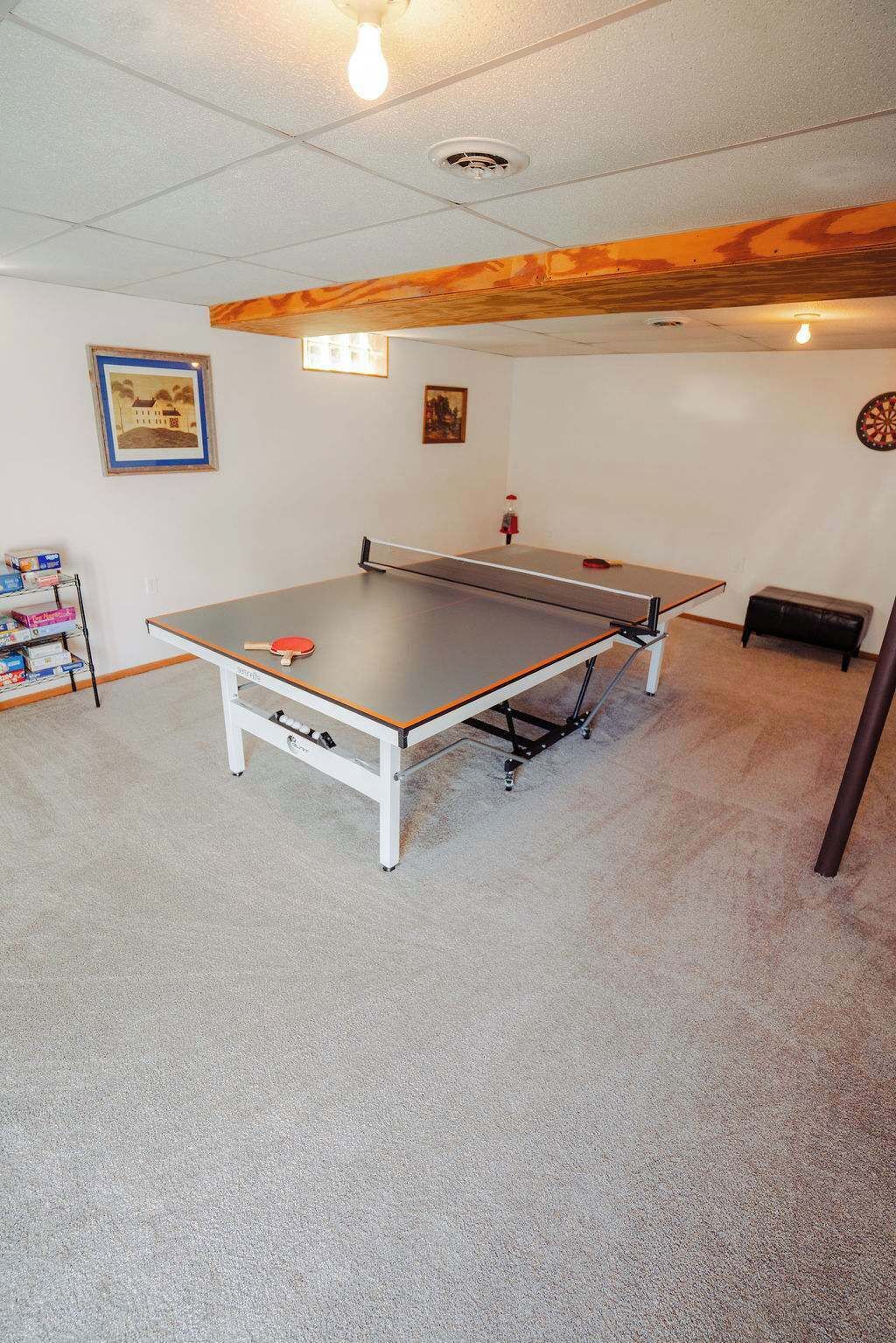 Una mesa de ping pong en una habitación con piso alfombrado, una estantería con cajas y un dardo en la pared.
