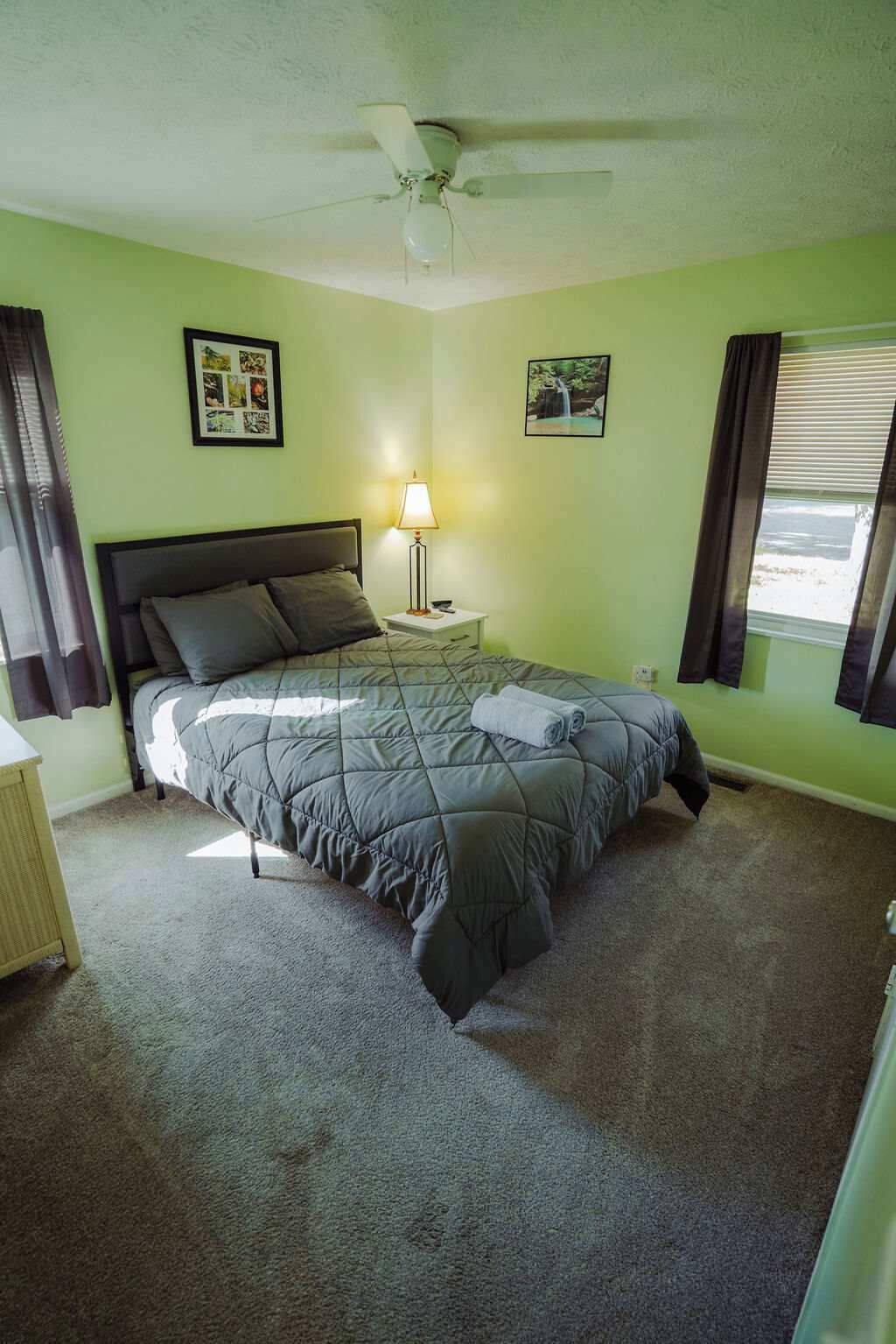Un dormitorio con cama, mesita de noche, lámpara, ventilador de techo y ventana con cortinas.
