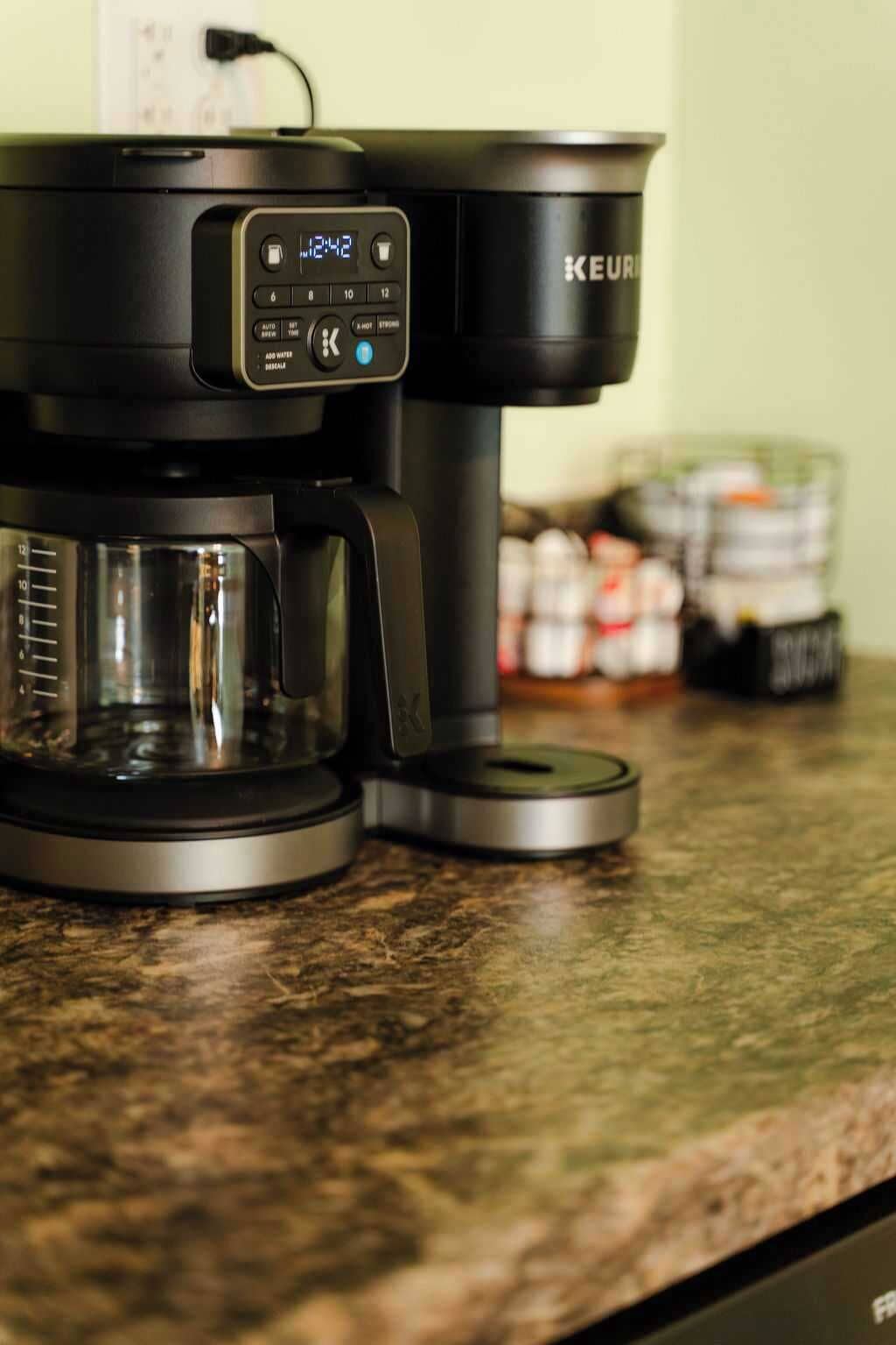 En Keurig kaffemaskine på en køkkenbordplade med en kande og en beholder med kaffekapsler i baggrunden.