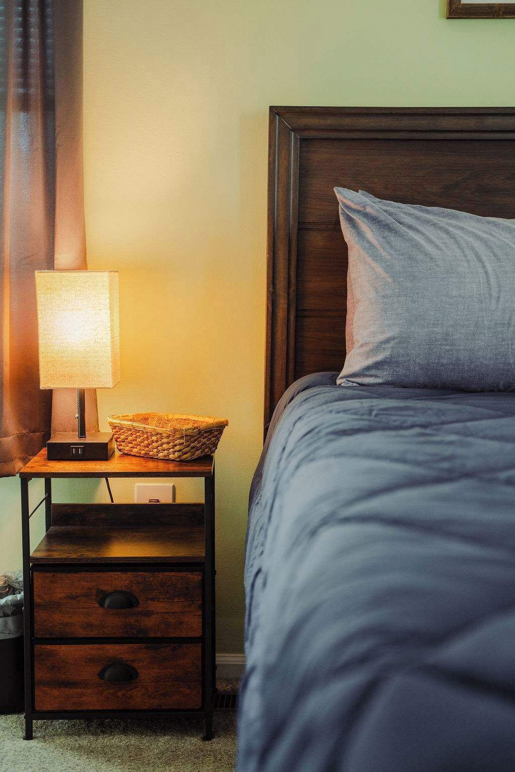 A neatly made bed with a grey pillow and a wooden nightstand beside it. A lamp with a white shade and a woven basket are also on the nightstand.