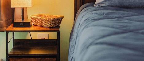 A neatly made bed with a grey pillow and a wooden nightstand beside it. A lamp with a white shade and a woven basket are also on the nightstand.