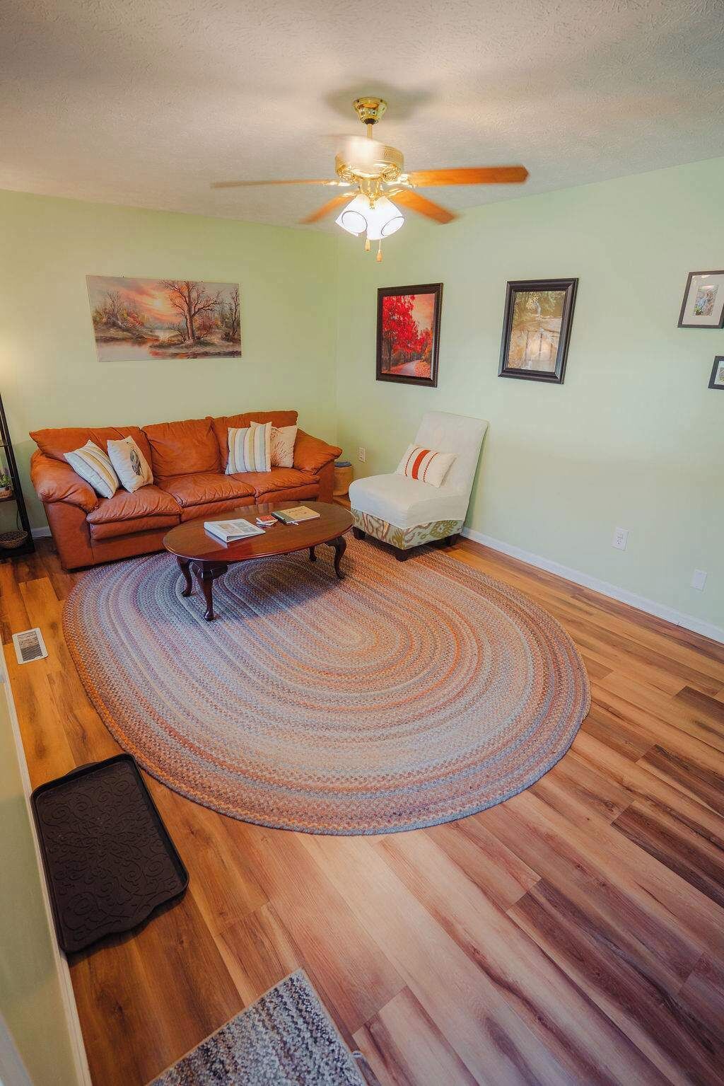 A living room with a wooden floor, a brown sofa, a white armchair, a coffee table, and a ceiling fan.