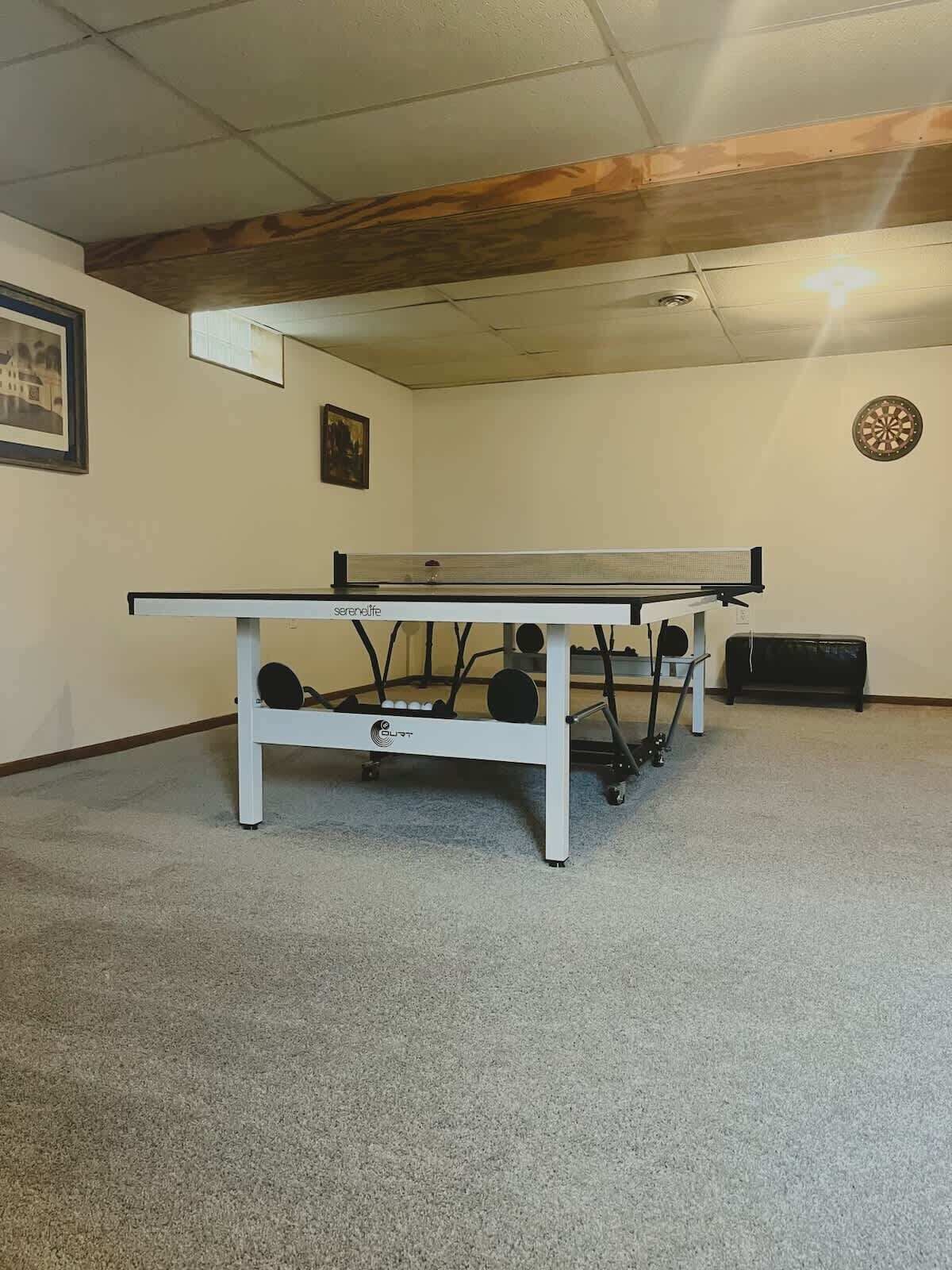 Une table de ping-pong dans une pièce dont le plafond est en bois, avec un jeu de fléchettes et un tableau encadré accroché au mur.