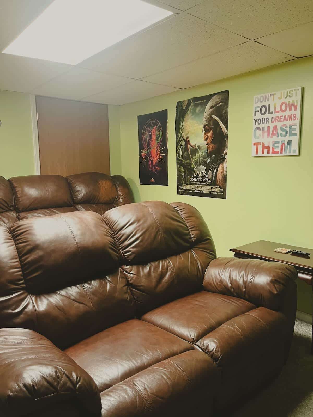 A room with a large brown leather couch, a wooden door, and two posters on the wall.