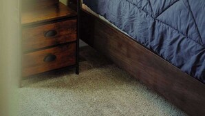 A neatly made bed with a dark blue quilt, a wooden nightstand with a basket, and a bedside lamp. - Blue Heron Cottage With Game Room, Deck & Hot Tub (Logan)