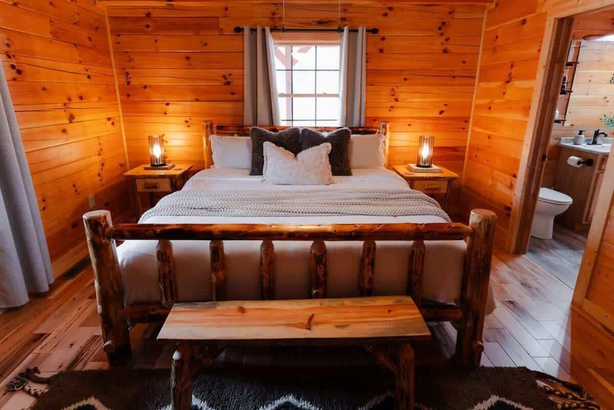 Puisesta hirrestä rakennettu mökin makuuhuone, jossa on sänky, yöpöyd ät, penkki ja ikkuna.