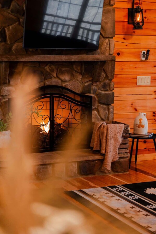 A cozy fireplace with a stone surround, a television mounted above it, and a rustic wooden wall. - Sleeps 16! Luxury Cabin With Game Room & Hot Tub (Logan)