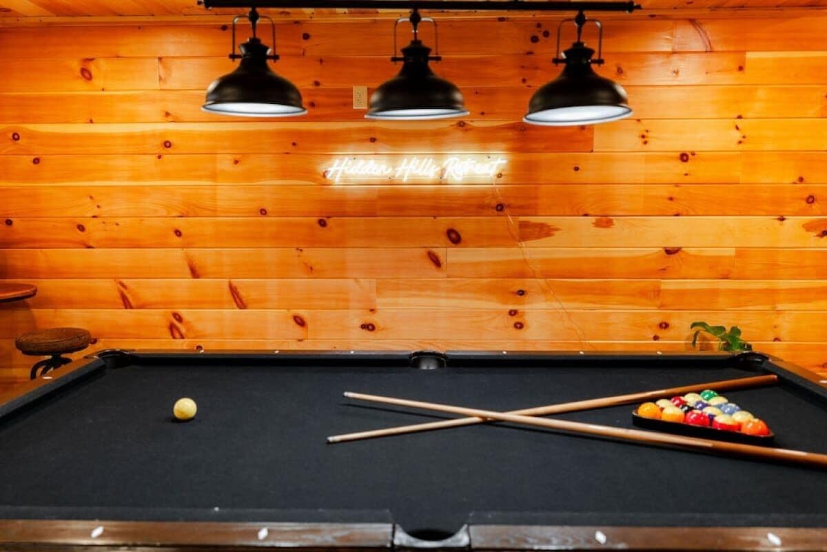 A pool table with a cue ball and a set of pool cues, three hanging lamps, and a neon sign on the wall.