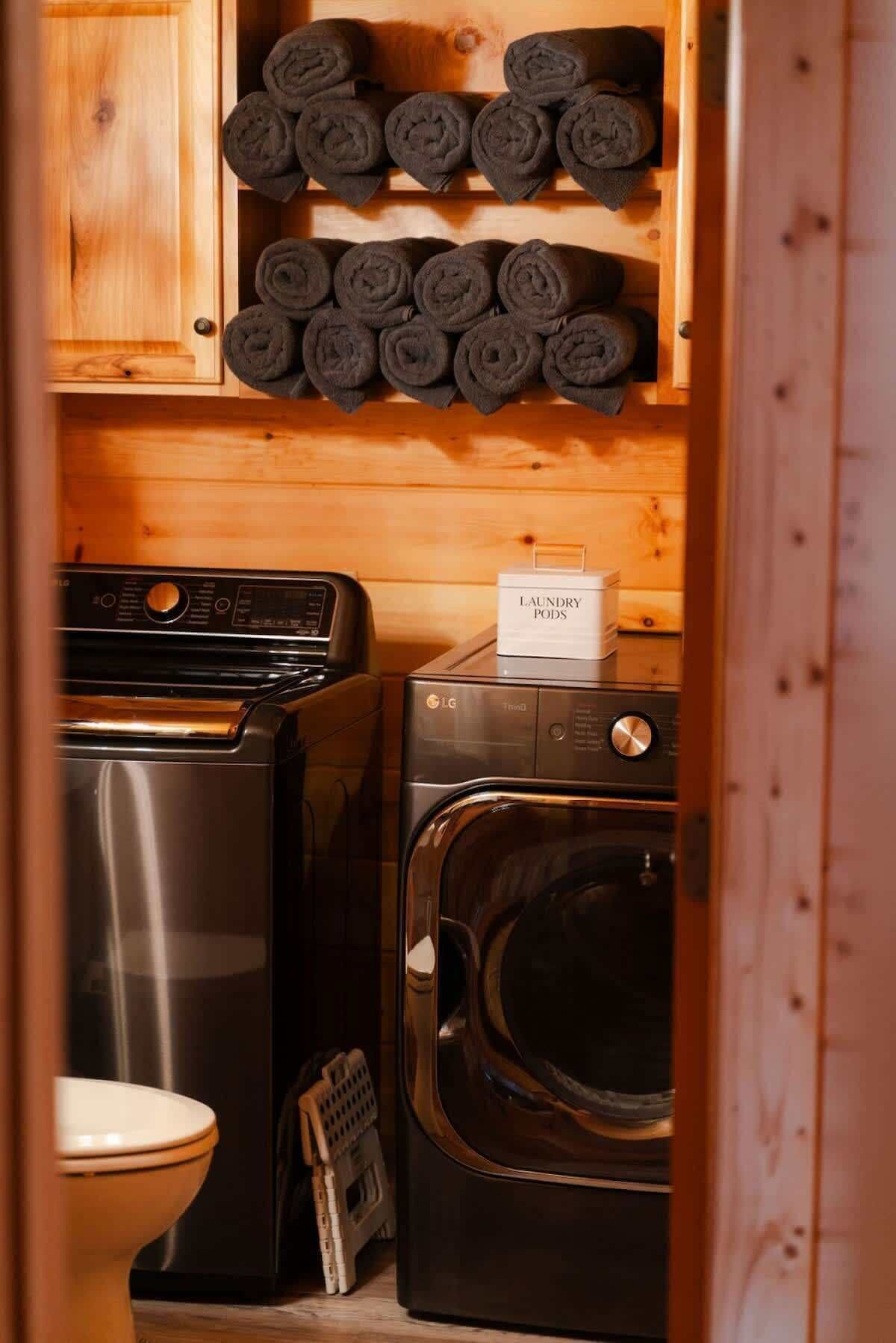A laundry room with a stacked washer and dryer, a rack of rolled towels, and a sign indicating laundry pods.