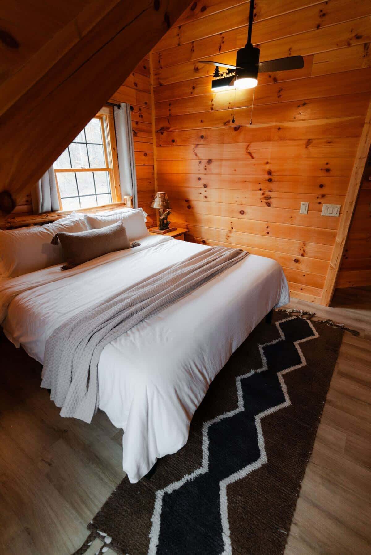 A cozy bedroom with a wooden ceiling, a bed with white bedding, a black and white rug, and a small table with a lamp.