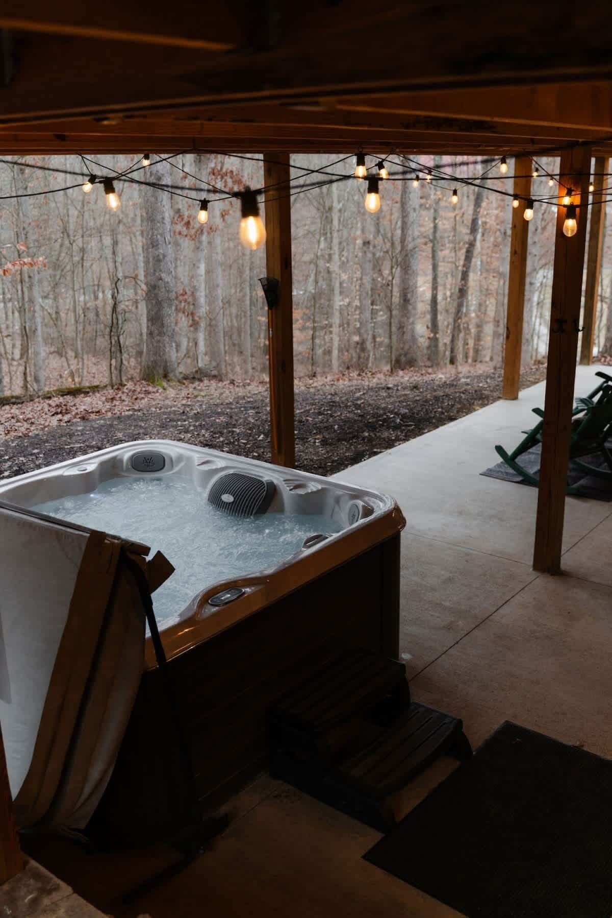 A hot tub under a wooden pergola with string lights, surrounded by a forested area.