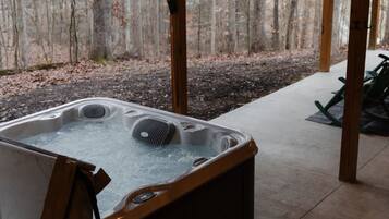 A hot tub under a wooden pergola with string lights, surrounded by a forested area.