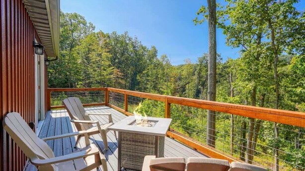 A wooden deck with outdoor seating and a fire pit, surrounded by a wooden railing and overlooking a forested area. - Tranquil Heights Cabin In Hocking Hills- HOT Tub! (Ray)
