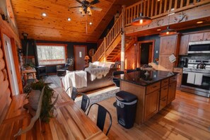 A rustic cabin interior with a wooden dining area, a cozy living room with a fireplace, a kitchen with modern appliances, and a staircase leading to an upper level. - Cozy Cabin With Hot Tub - Near all in Hocking Hills (Logan)