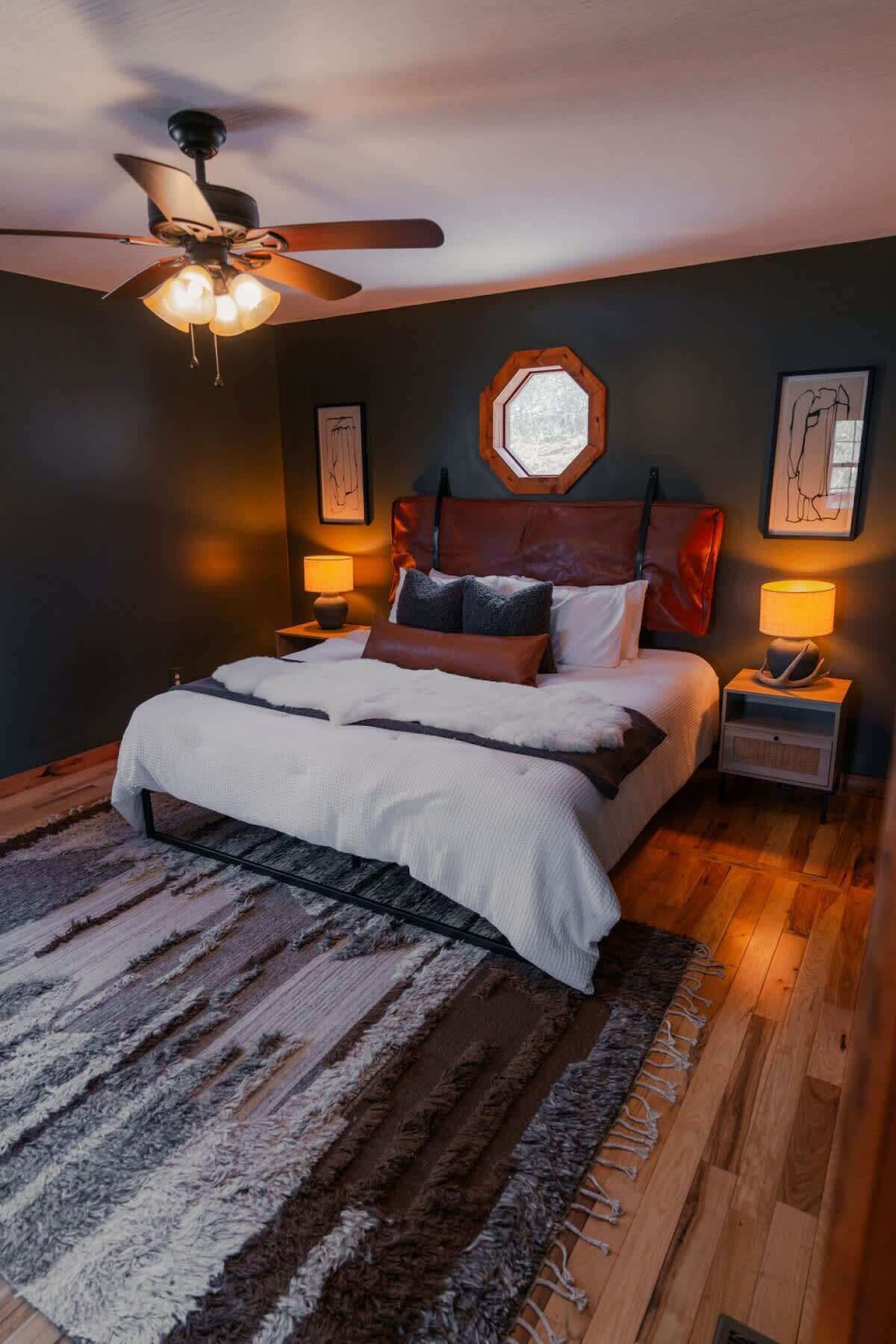 A bedroom with a bed, a ceiling fan, a window, and a nightstand with a lamp.