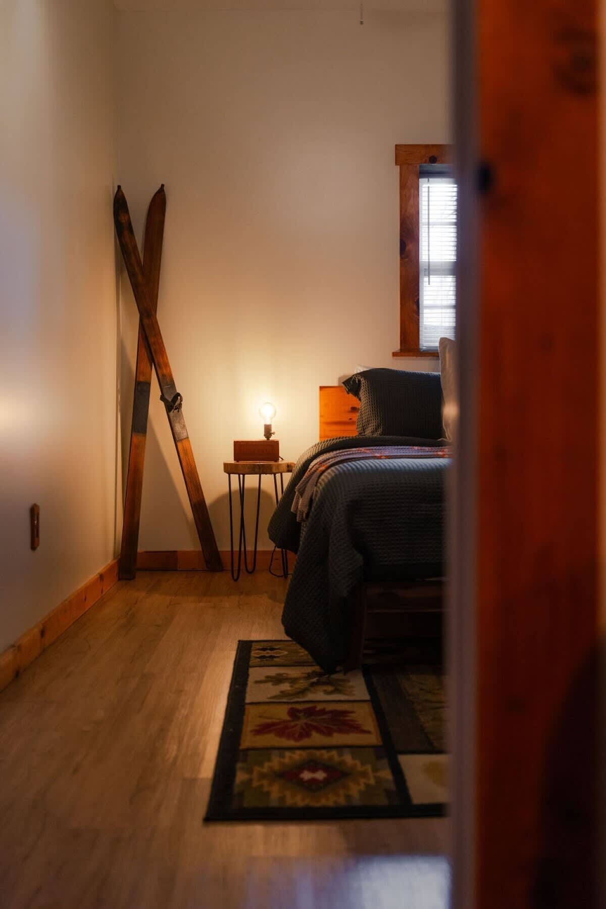 Un dormitorio con cama, piso de madera, una mesita con lámpara y una ventana.