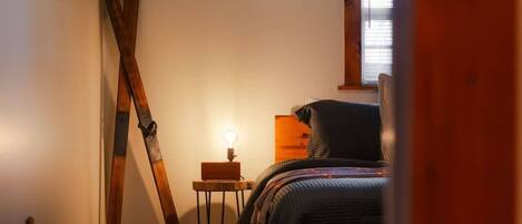 Un dormitorio con cama, piso de madera, una mesita con lámpara y una ventana.