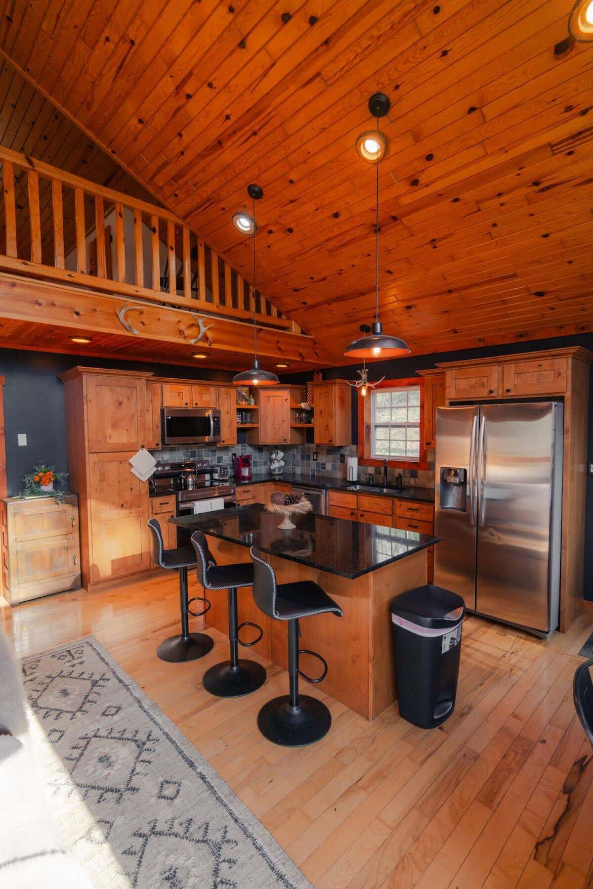 A spacious kitchen with wooden cabinets, a central island with bar stools, a stainless steel refrigerator, and a hanging light fixture.