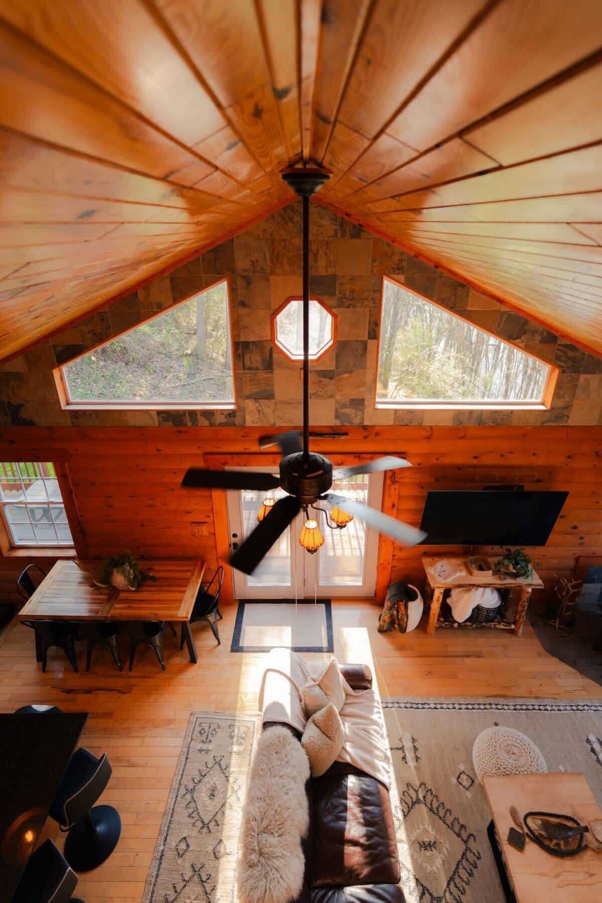 A cozy cabin interior with a sloped ceiling, wooden walls, a ceiling fan, a fireplace, and large windows.