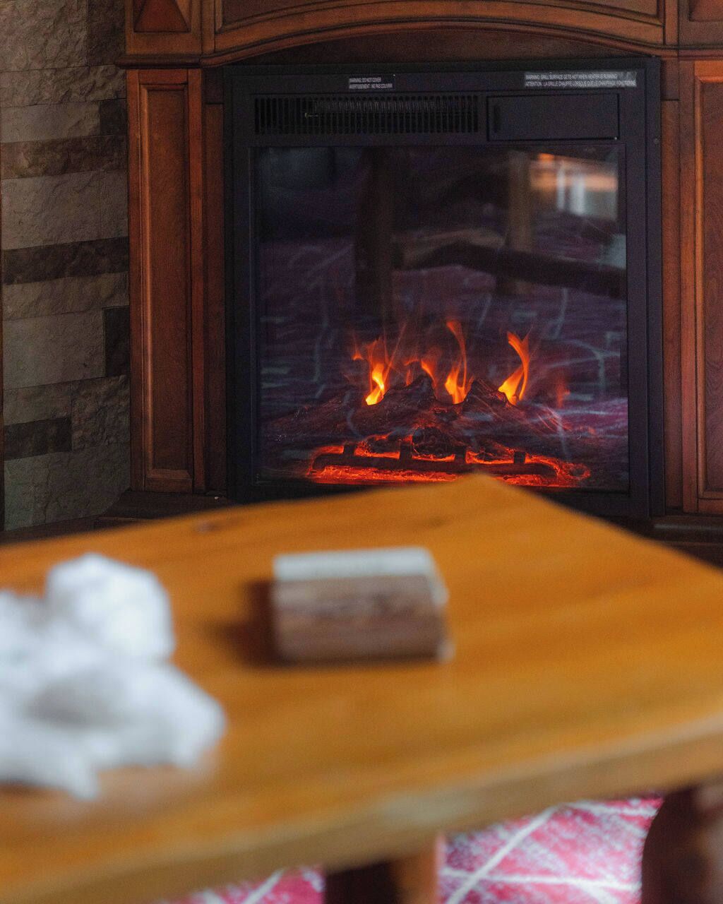 A fireplace with burning logs, a wooden table in the foreground, and a stone wall.