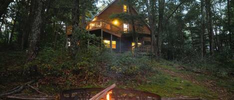 A cabin surrounded by forest, with a fire pit in the foreground.