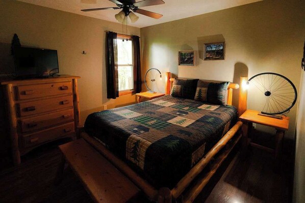 Une chambre à coucher comprenant un lit, une commode en bois, un ventilateur de plafond, une fenêtre avec des rideaux et une lampe fixée au mur.