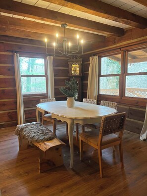 A wooden cabin interior with a round dining table, four chairs, a bench, a chandelier, and a large window with curtains. - Outdoors Sleeps7 Family Getaway Fun Arcade (Rockbridge)