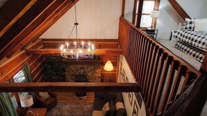 A cozy loft bedroom with a sloped ceiling, a stone fireplace, a chandelier, and a bed with checkered bedding. - Outdoors Sleeps7 Family Getaway Fun Arcade (Rockbridge)