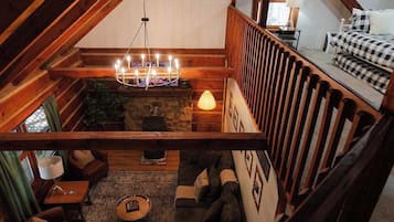 A cozy loft bedroom with a sloped ceiling, a stone fireplace, a chandelier, and a bed with checkered bedding.
