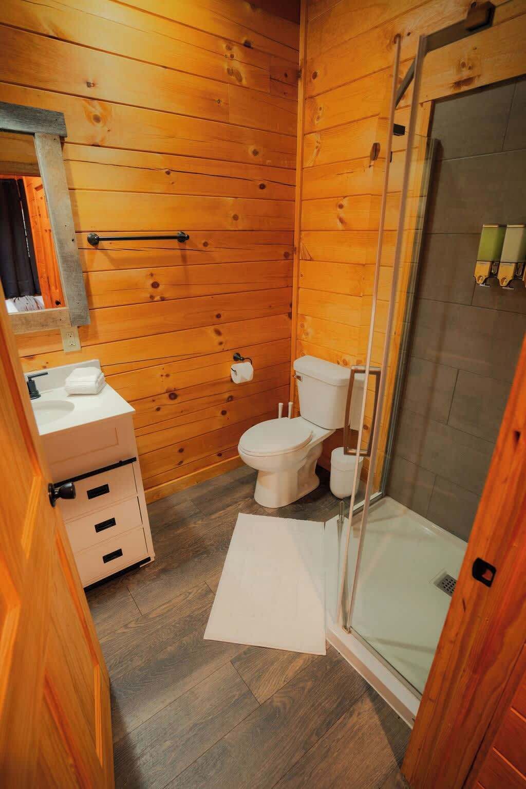 A compact wooden bathroom with a toilet, sink, and glass shower enclosure.