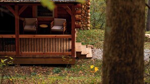 Una cabaña con terraza de madera y dos sillas, rodeada de árboles y hojas secas.