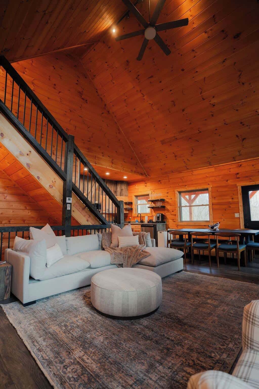 A spacious living room with a large wooden ceiling, a staircase, a dining area, and a kitchen in the background.