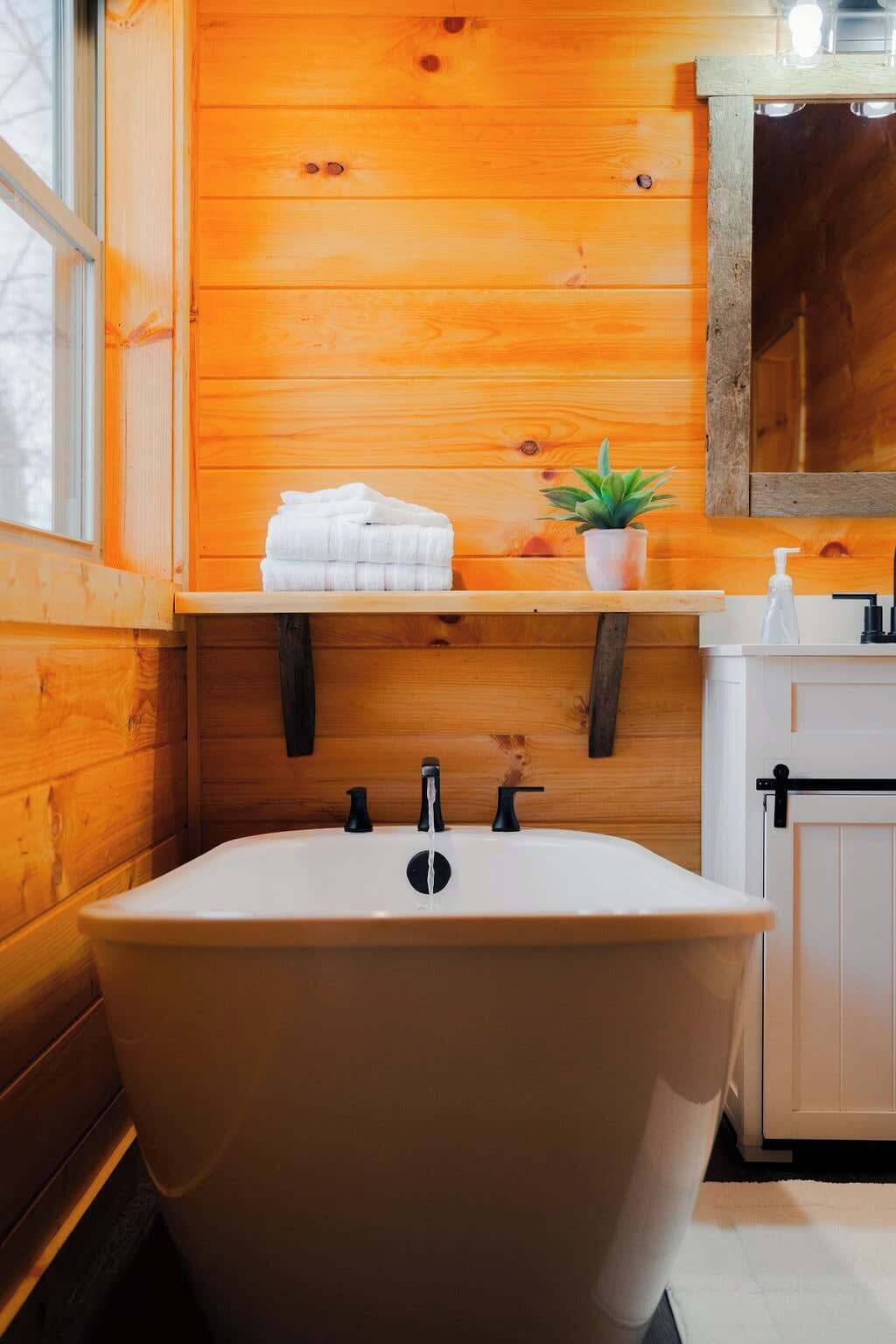 Une salle de bain avec une baignoire sur pieds, des murs en bois, une étagère avec des serviettes et une plante en pot, ainsi qu’un lavabo blanc avec un miroir au-dessus.