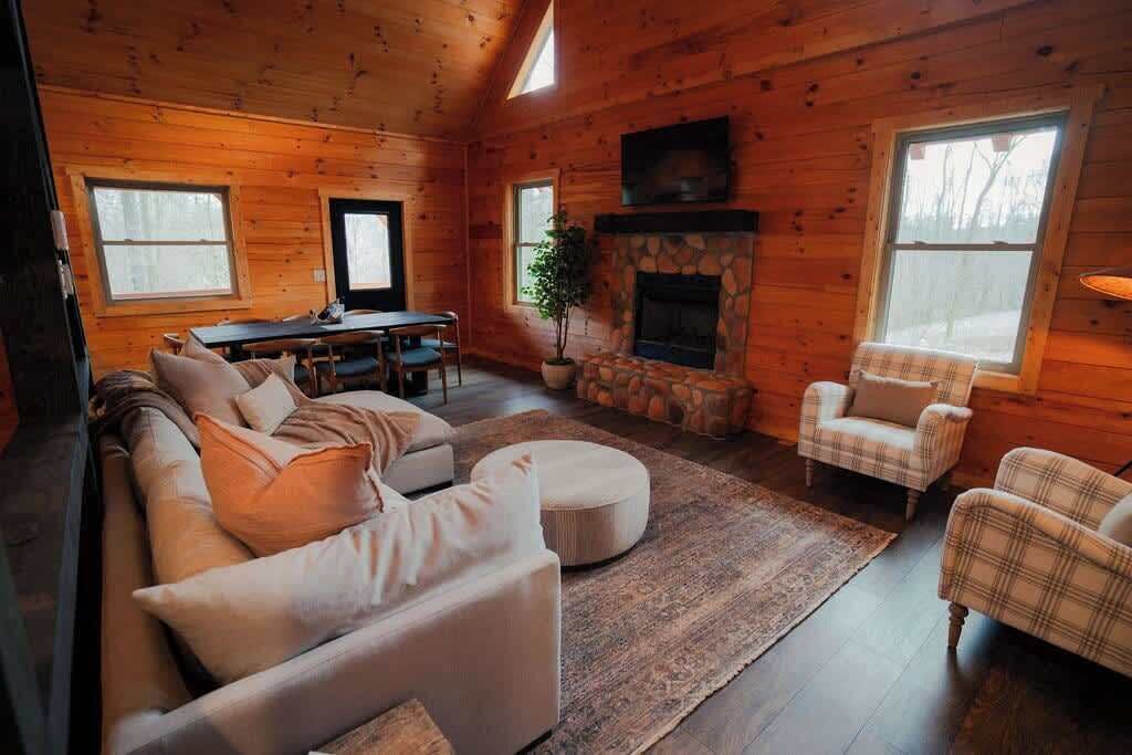 A cozy cabin interior with a stone fireplace, a sofa, a plaid armchair, and a dining area.