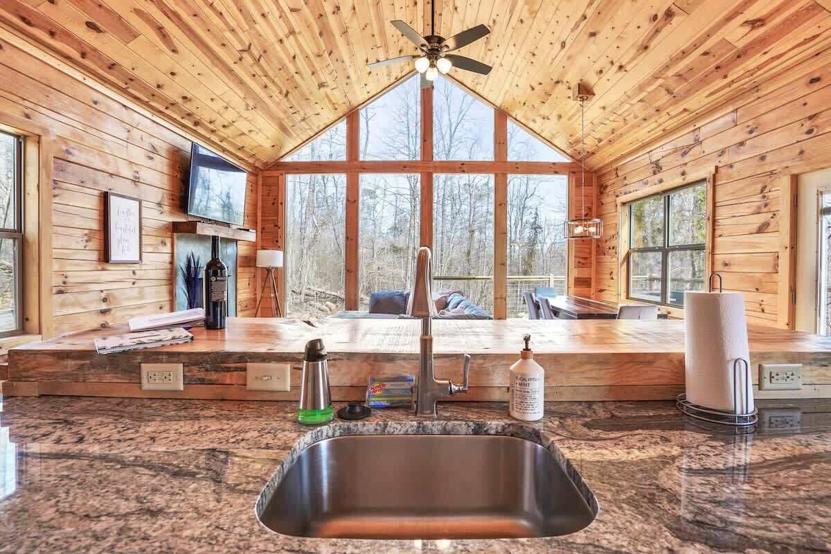 A wooden cabin interior with a kitchen area, large windows, and a ceiling fan.