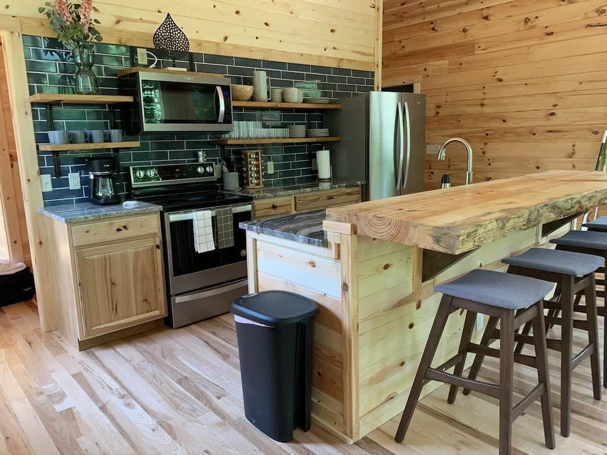 A modern kitchen with wooden cabinets, a built-in oven, a microwave, and a refrigerator. There are bar stools and a trash can.
