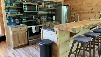 A modern kitchen with wooden cabinets, a built-in oven, a microwave, and a refrigerator. There are bar stools and a trash can.