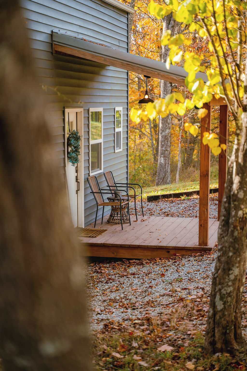Eine Holzterrasse mit zwei Korbstühlen und einem kleinen Tisch, eine Tür mit einem Kranz und Blick auf Herbstbäume.