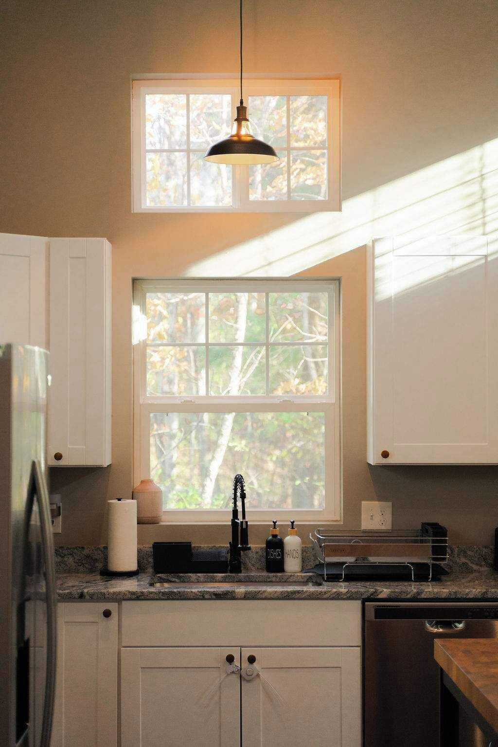 A kitchen with white cabinets, a window with a view of trees, a hanging pendant light, and a countertop with a sink and dish soap.
