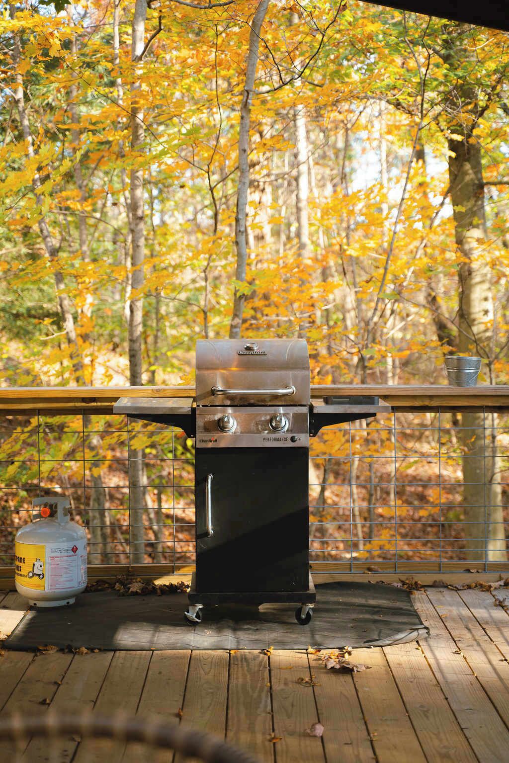 Un barbecue portable sur une terrasse en bois dotée d’une rambarde métallique, entouré d’un feuillage automnal.