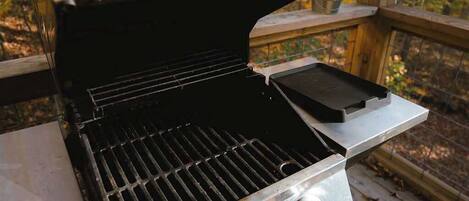 A portable grill with a metal handle and two knobs, placed on a wooden deck with a view of autumn trees.