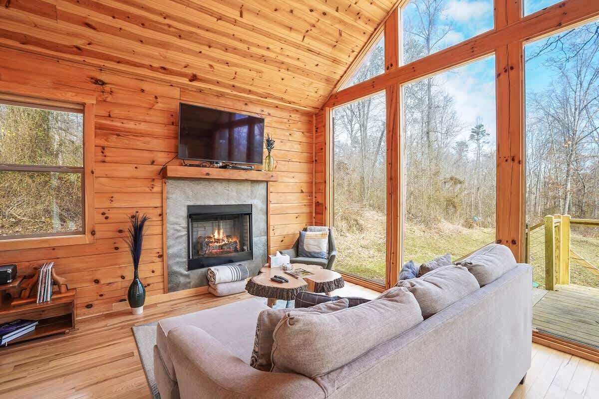 Interior acolhedor de uma cabana com lareira, uma televisão montada acima dela, um sofá e uma janela grande com vista para a floresta ao redor.