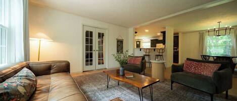 A living room with a sofa, a coffee table, and a couch. There is a door leading to a kitchen area.