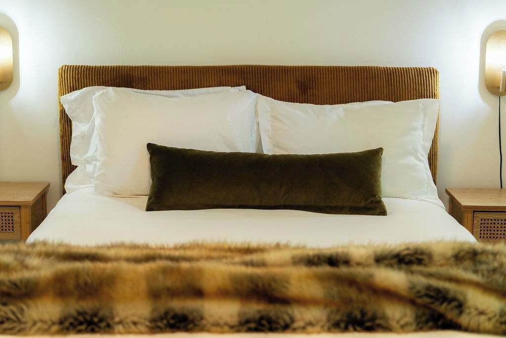 A bed with white linens and a dark brown pillow, flanked by two bedside tables with lamps.