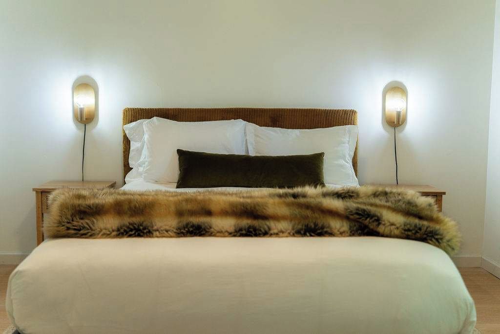 A bed with a white mattress, a dark brown pillow, and a fur throw. There are two bedside tables with lamps on either side of the bed.