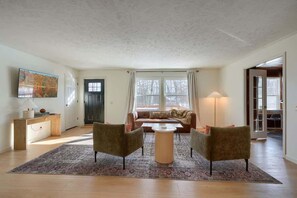 Een woonkamer met een bank, twee fauteuils, een salontafel en een vloerkleed. Er hangt een schilderij aan de muur, en er staat een lamp. Een deur leidt naar een andere kamer.
