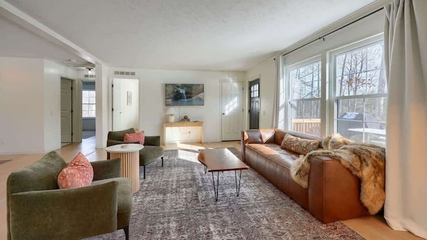 A spacious living room with a large window, a brown leather sofa, a green armchair, and a small round coffee table.