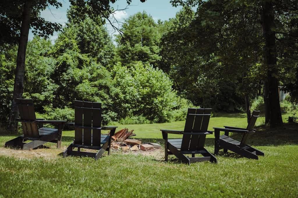 A row of Adirondack chairs facing a fire pit in a grassy area with trees in the background.