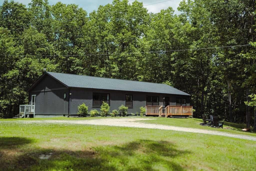 A single-story building with a dark roof and a porch, surrounded by a grassy area and dense trees.