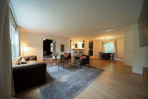 Un salón amplio con un sofá, sillones y una mesa de centro. Hay un comedor con una mesa y sillas. La habitación tiene suelo de madera y una alfombra grande.